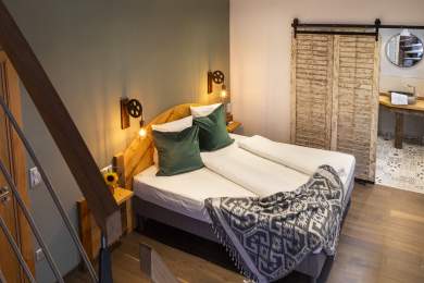 Chambre avec deux lits simples côte à côte, murs en pierre apparente, coussins décoratifs à motifs rouges, lampes murales noires et fenêtre avec rideaux gris