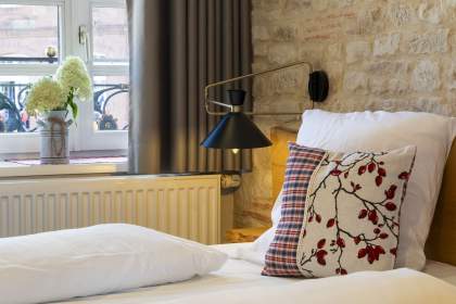 Détail d’une chambre avec lit, coussin décoratif à motifs rouges, lampe murale noire orientable, mur en pierre apparente et fenêtre avec rideaux gris et vase de fleurs blanches