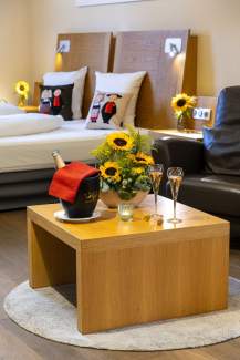 Table basse en bois avec un seau à champagne, deux flûtes et un bouquet de tournesols, dans une chambre moderne avec lit double et décorations florales.