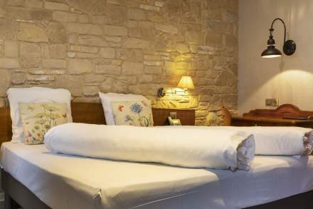 Chambre avec lit double, mur en pierre apparente, deux oreillers décorés de motifs floraux, lampe murale allumée et bureau en bois avec téléphone