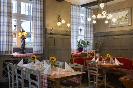 Salle de restaurant traditionnelle avec nappes à carreaux rouges et blancs, tables dressées avec serviettes pliées, bouquets de tournesols, chaises en bois gris, banquettes rouges, lampes suspendues et décoration rustique près de fenêtres à rideaux à carreaux.