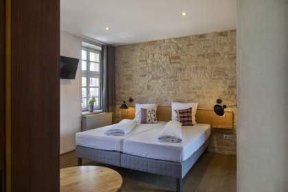 Chambre avec deux lits simples côte à côte, mur en pierre apparente, coussins décoratifs, lampes murales noires et fenêtre apportant lumière naturelle