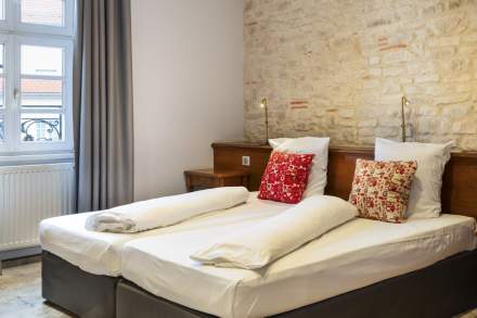 Chambre avec deux lits simples côte à côte, tête de lit en bois, mur en pierre apparente, coussins rouges à motifs, et fenêtre avec rideaux gris