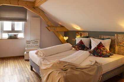 Chambre cosy sous les combles avec lit double, coussins à motifs floraux, poutres en bois apparentes et lumière douce